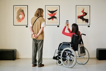 Two people in modern gallery observing and photographing abstract art pieces displayed on wall. One...