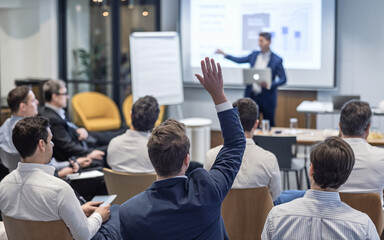 Group of participants in an interactive business seminar, with one individual raising their hand while seated and another person presenting in the background with a laptop.

