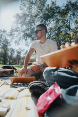 A young man sitting on a picnic blanket in a sunny park, wearing sunglasses and enjoying a relaxed conversation during a casual outdoor gathering with friends.