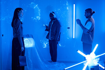 Participants engaging with futuristic techno art installation illuminated by blue lighting. Diverse group examining interactive displays with immersive surroundings