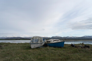 Abandoned derelict boats on a grassy shoreline with water and snowy mountains in the background.