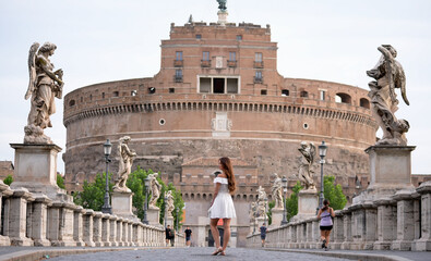 A stylish woman in a white dress strolls across Ponte Sant Angelo, framed by angel statues and the...