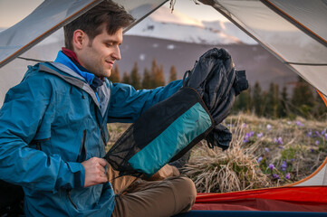 Young male hiker unpacking a sleeping bag from a compression sack in his tent at sunset in the...