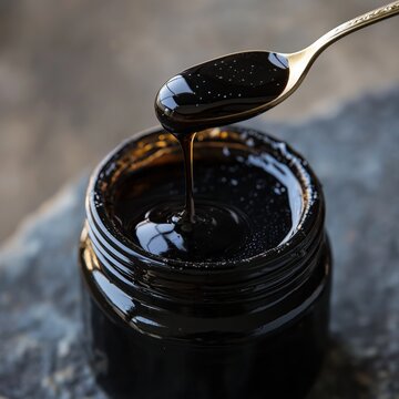 Open jar of black shilajit honey drips from spoon on natural stone backdrop. Superfood health product. Natural remedy. Healthy lifestyle. Dark rich honey. Natural ingredient. High quality food.