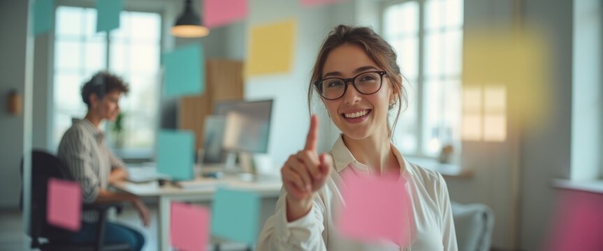 In a vibrant office setting, a cheerful young woman with glasses playfully points at colorful sticky notes, embodying creativity and collaboration. Behind her, a focused colleague works diligently at - Powered by Adobe