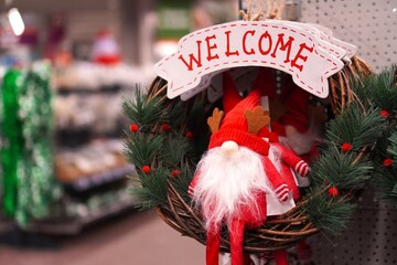 Soft Christmas figurine with gnome on a wreath of branches with the inscription Welcome on the background of shelves with Christmas decorations in the store before the winter holidays