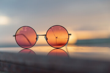 Pink sunglasses with cracked glass lie on the parapet of the promenade against the setting sun. The concept of a day, a year, a life coming to an end with the realization that there was a deception.