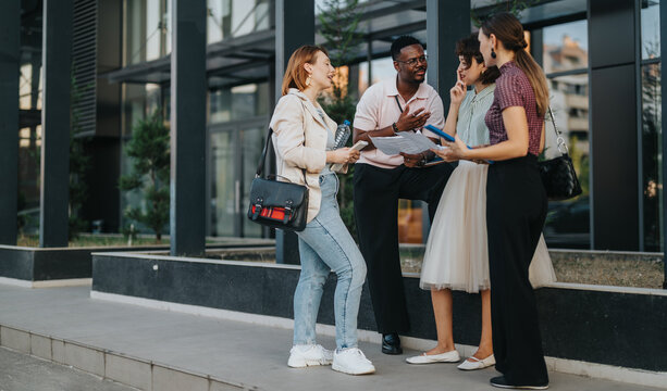 A group of young business people engage in a lively discussion outside a contemporary office building, holding documents and digital devices. This image captures teamwork and collaboration in an urban