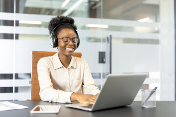 Young professional woman wearing headphones working on laptop in modern office. Expression of productivity and focus, with smartphone nearby indicating multitasking capability.