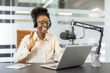 Joyful woman in headphones recording podcast in office setting with microphone, laptop, and tablet....