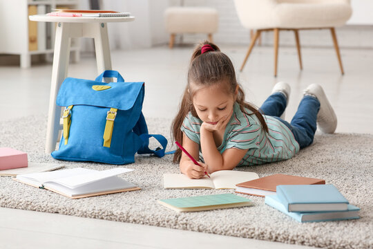 Cute little girl with notebooks, backpack and pencil lying on floor and studying at home - Powered by Adobe