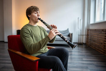 Clarinet player. Concentrated clarinetist playing flute woodwind music instrument sitting on armchair at home. Musical instruments. Man plays the oboe in living room. Relaxed mental hobby concept © DimaBerlin