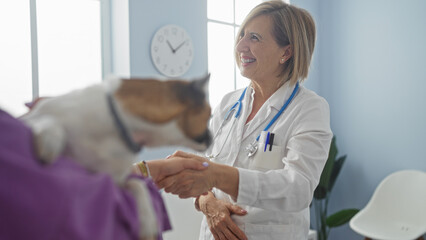 A middle-aged blonde woman vet smiles while shaking hands with an adult owner at a veterinary...