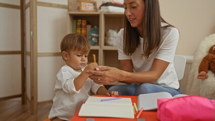 Woman helping son write indoors in a kindergarten playroom, symbolizing family love and education in a warm, nurturing environment.