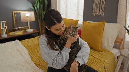 A young hispanic woman cuddles her kitten while sitting on a cozy bed in a warmly decorated bedroom...