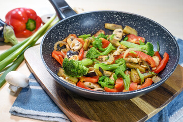 Colorful mixed vegetables in a frying pan such as broccoli, bell pepper, mushrooms, eggplant and onions on a wooden kitchen board, healthy vegetarian meal