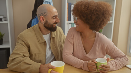 A cheerful interracial couple enjoys a cozy coffee break together indoors, sharing a laugh in a casual home setting.