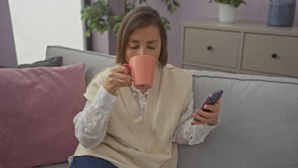 Adult woman sips from mug while browsing smartphone in a cozy living room setting, conveying...