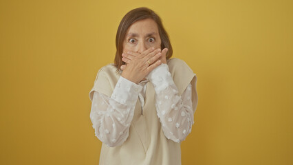 Mature woman covering mouth with hands in surprise or silence against a yellow background