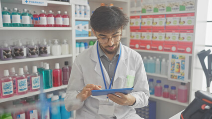 A young man with a beard in a white lab coat analyzes data on a tablet in a well-stocked pharmacy.