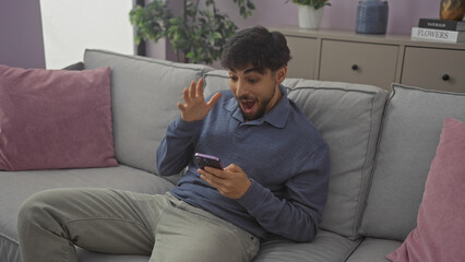 Fototapeta premium Excited man with beard in modern apartment living room using smartphone on couch