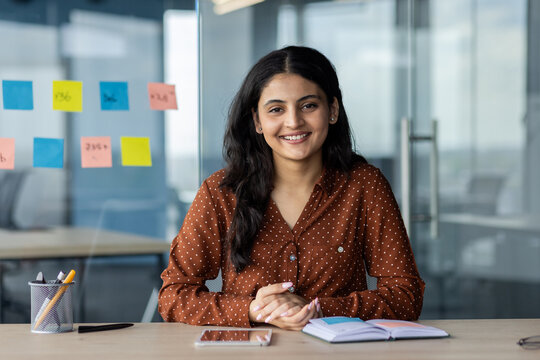 Latin American businesswoman smiling at desk in modern office. Confident, professional ambiance with smartphone, notebook. Represents success, motivation, casual work environment.