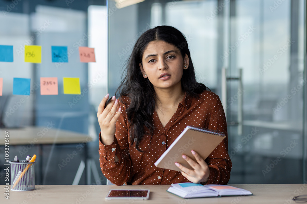 Canvas Prints Latin American businesswoman holding notebook, engaged in conversation in modern office. Emphasizes communication skills with phone and sticky notes on table, illustrating professional setting.