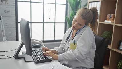 Young blonde woman, a doctor, multitasks on a computer while making a phone call in a bright hospital office