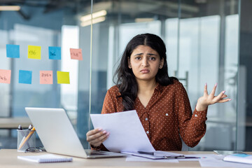 Latin American woman businesswoman looks confused while reviewing documents in modern office. Paperwork challenges create frustration. Laptop and sticky notes visible.