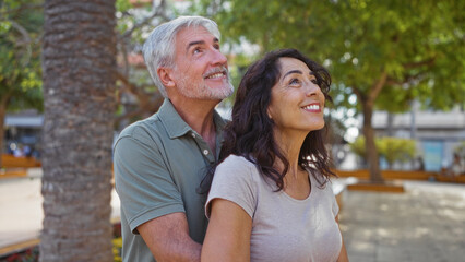 Obraz premium Middle-aged couple smiling happily in a sunny urban park setting, with man embracing woman amidst lush greenery, capturing the essence of love and togetherness outdoors.