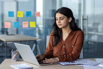 Latin American woman businesswoman working on laptop in office with glass walls and sticky notes....