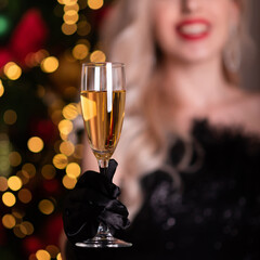 Beautiful girl in a black evening dress and black gloves against the background of a Christmas tree. Holds a glass of champagne in his hand and smiles. Focus on the glass. Celebrating Christmas