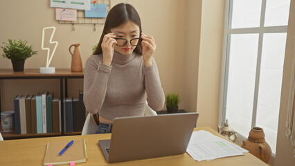 A young asian woman adjusts her glasses while working on a laptop in a modern home office setup.