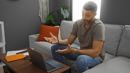 Man communicating online in home living room, sitting on a comfortable sofa with laptop, engaged in a video call, expressing thoughts through gestures.