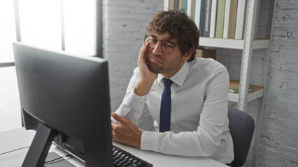 Man looking tired in office with computer wearing glasses and white shirt beside shelves with books in bright indoor workspace during workday.