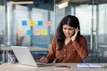 Latin American woman businesswoman at desk with laptop, phone experiencing ear pain. Expression...