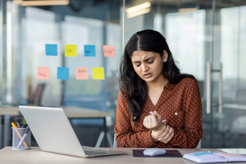 Latin American businesswoman sitting at office desk with laptop experiencing wrist pain. Shows...