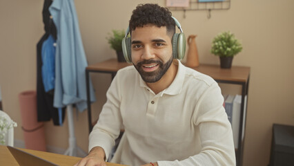 A smiling young hispanic man with headphones sits at an indoor home office desk, exuding casual professionalism and comfort.