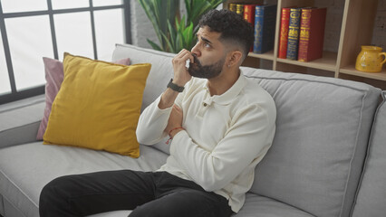 A thoughtful young hispanic man with a beard sits on a gray sofa in a modern living room, poignant and contemplative