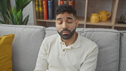 Obraz premium Handsome young hispanic man with beard sitting thoughtfully on gray couch in cozy living room apartment.