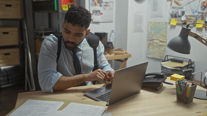 A focused hispanic man investigates in a cluttered detective office, paperworking at his laptop...