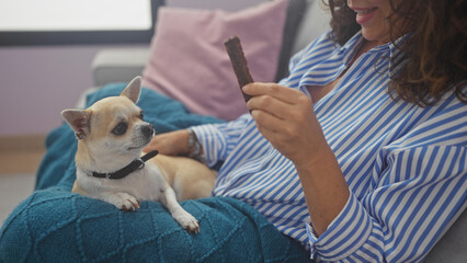 A middle-aged hispanic woman in a striped shirt smiles at her chihuahua indoors, holding a treat in a cozy room.