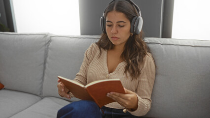 Woman listening to music on headphones while reading a book indoors sitting on a couch in a cozy living room.
