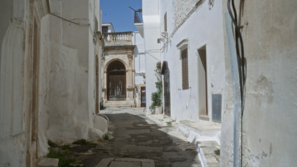 Obraz premium A narrow, sunlit alleyway in ostuni, puglia, italy showcases charming whitewashed buildings, rustic architecture, and a serene, historic mediterranean ambiance.