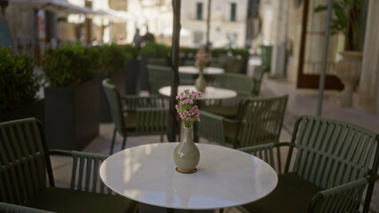 Outdoor cafe scene in puglia, southern italy, featuring a pink flower arrangement on a white table with green chairs, blurred alley view, and a distant walking man.