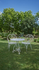 Two empty white wrought-iron chairs and a round table set on green grass in a lush garden under a clear blue sky in puglia, italy, europe, during daytime.