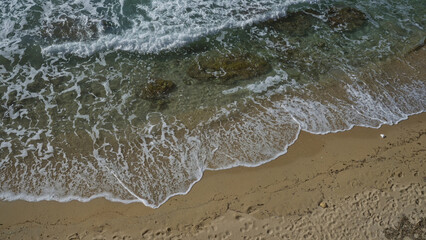 Aerial view of the clear turquoise waters and sandy shore of gallipoli, puglia, salento in italy, showcasing waves, rocks, and footprints on the beach.