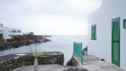Fototapeta premium Blurred coastal scene in lanzarote with white buildings, green shutters, and rocky shoreline along the sea under a cloudy sky