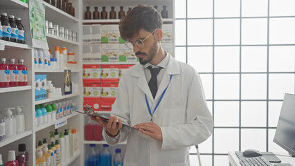 A young man pharmacist in a white coat inspects a clipboard within a modern drugstore interior.