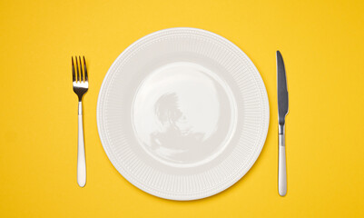 Round white empty ceramic plate and knife with fork on yellow background, table setting.